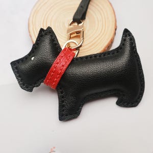 May include: A black leather dog-shaped keychain with a red collar and gold-colored hardware. The keychain is a stylized representation of a Scottish Terrier. The dog has a small white dot for an eye and is attached to a black leather strap.
