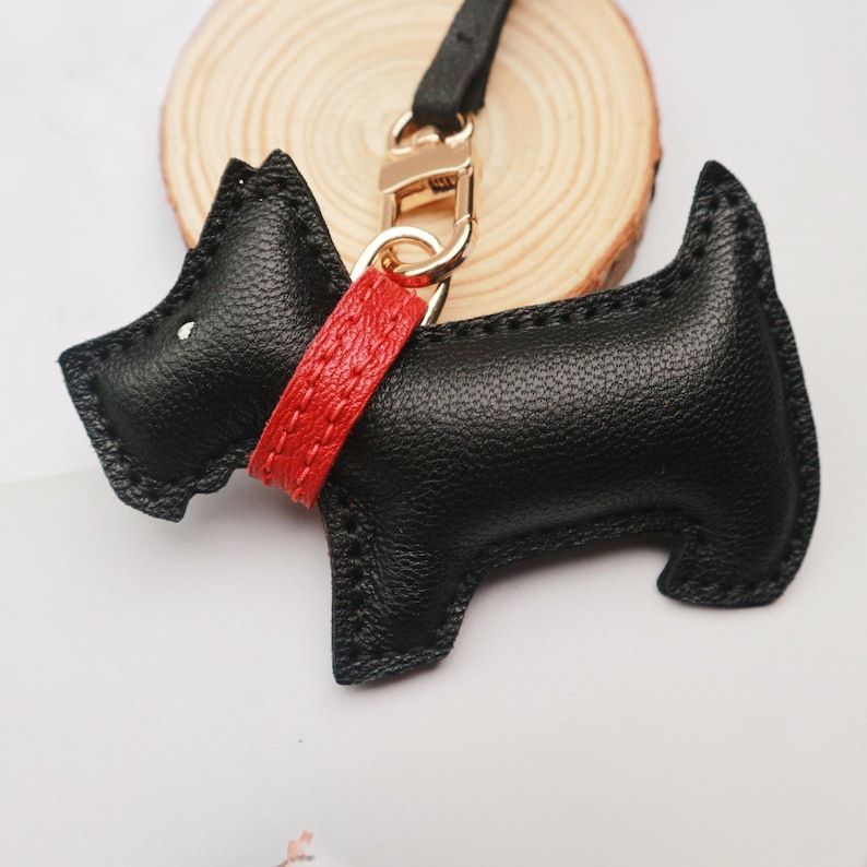 May include: A black leather dog-shaped keychain with a red collar and gold-colored hardware. The keychain is a stylized representation of a Scottish Terrier. The dog has a small white dot for an eye and is attached to a black leather strap.