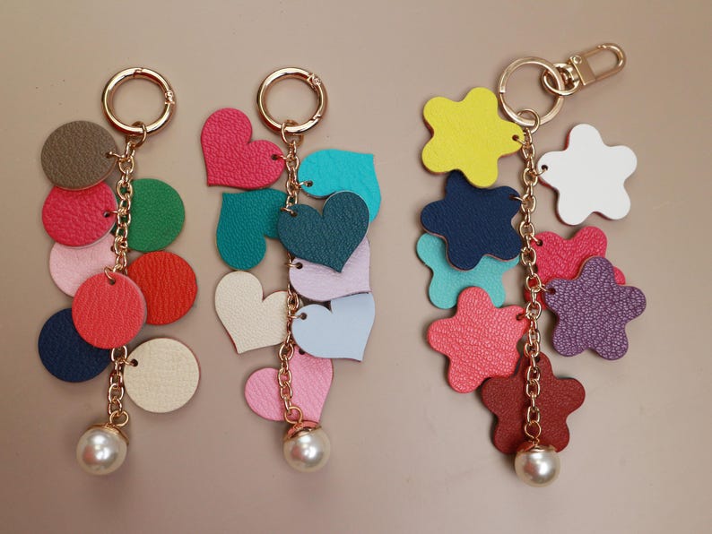 May include: Three colorful keychains with different shapes and colors. Each keychain has a gold chain and a pearl charm. The keychains feature hearts, stars, and circles in various colors, including pink, blue, green, red, and purple.