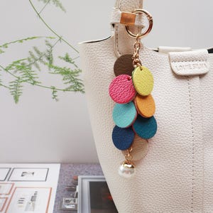 May include: A white handbag with a gold chain and a colorful leather tassel keychain. The keychain features a series of round leather pieces in shades of pink, yellow, blue, and green, with a pearl-like bead at the bottom.