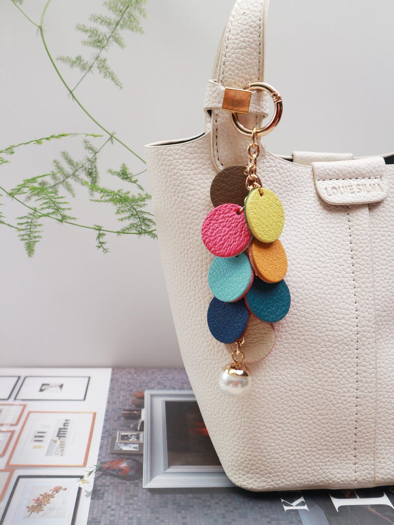 May include: A white handbag with a gold chain and a colorful leather tassel keychain. The keychain features a series of round leather pieces in shades of pink, yellow, blue, and green, with a pearl-like bead at the bottom.