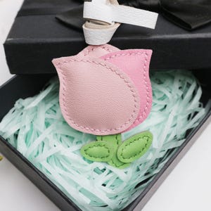 May include: A pink and light pink leather rose keychain with green leaves, presented in a black gift box with a white ribbon. The rose is made of stitched leather and has a detailed design.