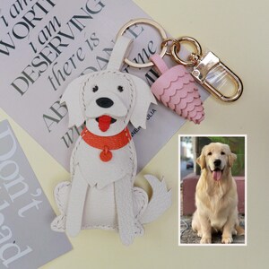 May include: White leather dog keychain with an orange collar and red tongue. The keychain has a gold-colored clasp and ring, with a pink, scalloped charm. A photo of a golden retriever is in the bottom right corner.