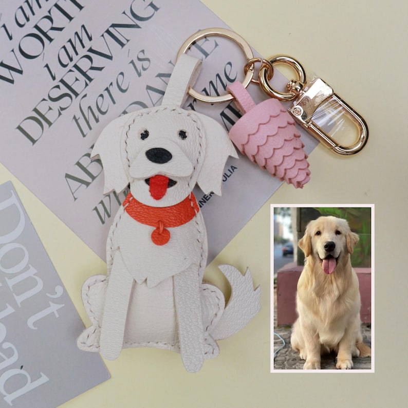 May include: White leather dog keychain with an orange collar and red tongue. The keychain has a gold-colored clasp and ring, with a pink, scalloped charm. A photo of a golden retriever is in the bottom right corner.