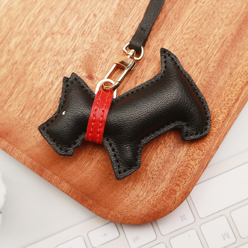 May include: A black leather dog-shaped keychain with a red collar and gold-colored clasp. The keychain is attached to a black strap. The dog has a white dot for an eye. The keychain is on a wooden surface.