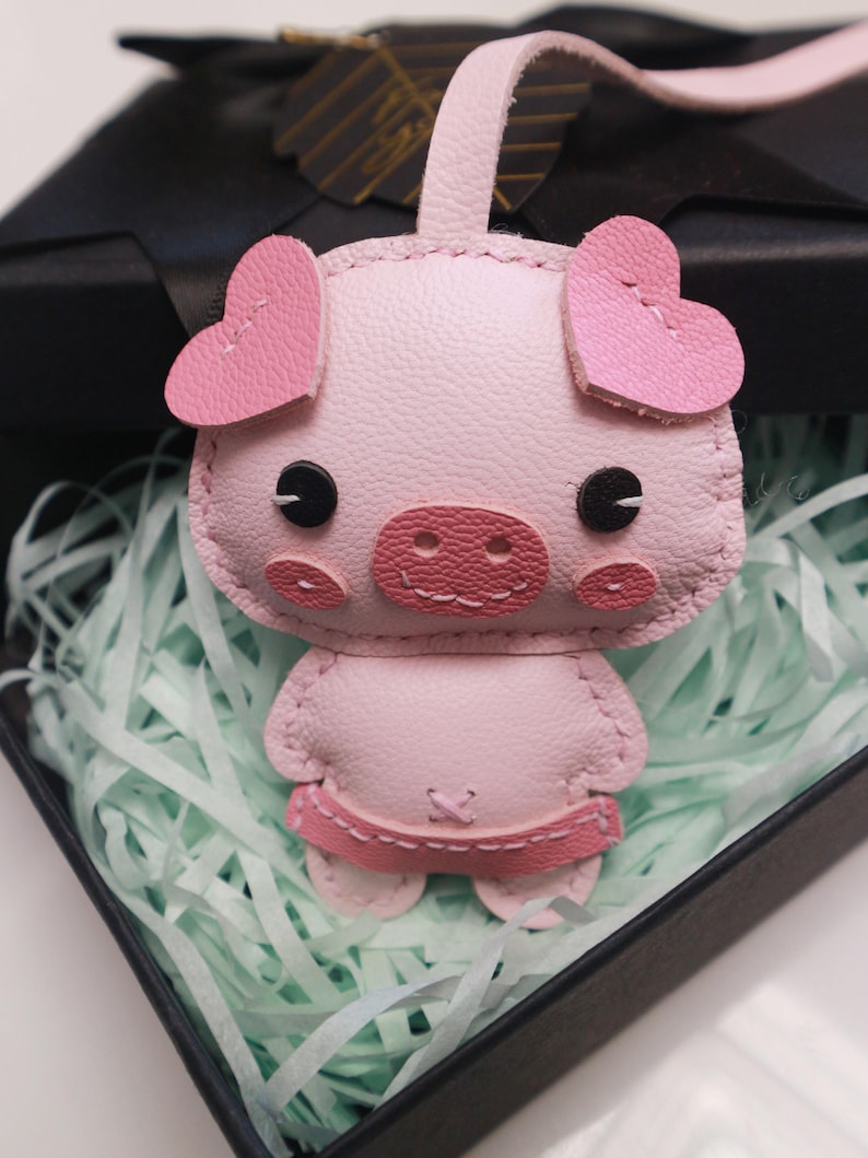 May include: A pink leather pig keychain with heart-shaped ears and black eyes. The pig is wearing a pink dress and has a stitched-on snout.