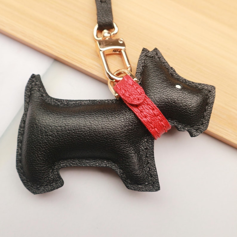 May include: A black leather dog-shaped keychain with a red collar. The keychain has a gold-colored clasp and a black strap. The dog has a textured surface and is a stylized representation of a terrier.