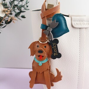 May include: A brown leather dog keychain with a blue collar and a red tongue, attached to a leather strap with a heart and bow charm. A bone-shaped tag reads "Rocky". The keychain hangs from a white handbag.