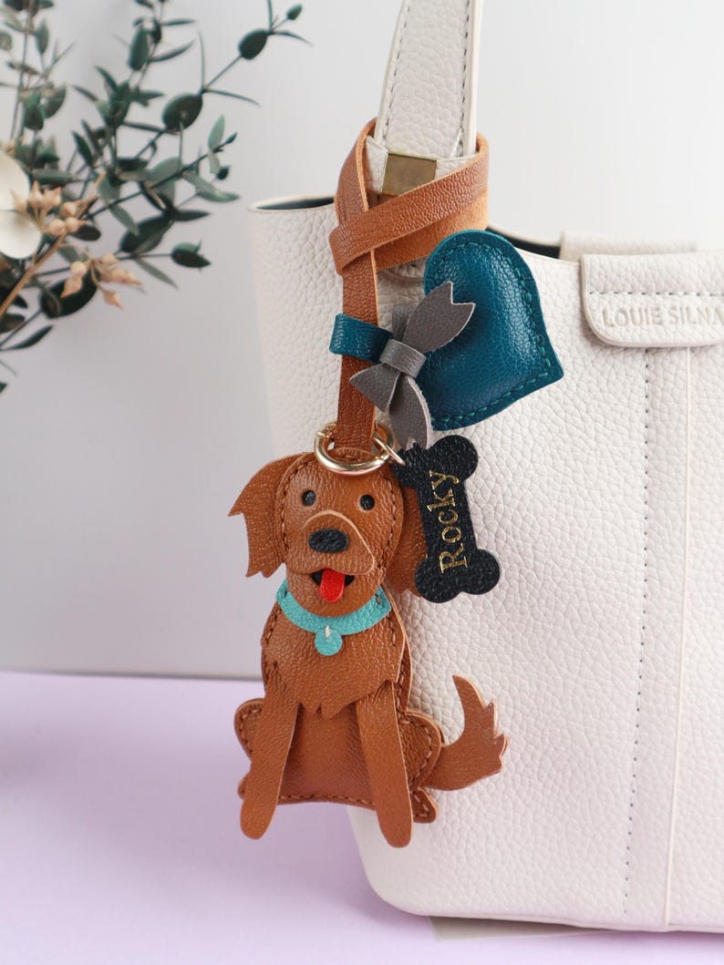 May include: A brown leather dog keychain with a blue collar and a red tongue, attached to a leather strap with a heart and bow charm. A bone-shaped tag reads "Rocky". The keychain hangs from a white handbag.