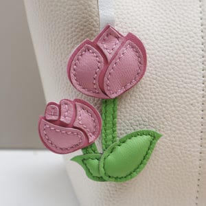 May include: A close-up of a white leather bag with a pink leather tulip flower applique. The applique features two tulips with green stems and leaves. The flowers are a light pink color with stitched details.