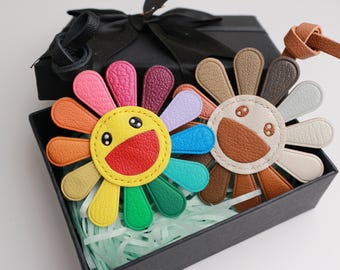 May include: Two colorful leather flower keychains with smiling faces. The flowers are made of different colored leather pieces and have a yellow center with a smiling face. The keychains are in a black gift box with white tissue paper.