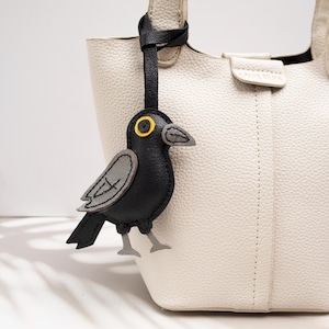 Peut inclure: Un sac à main texturé de couleur crème avec une lanière en cuir noir attachée à une breloque en forme d'oiseau noir et gris. La breloque oiseau présente un détail d'œil jaune, un bec gris et des pattes grises, suspendue à la poignée du sac.