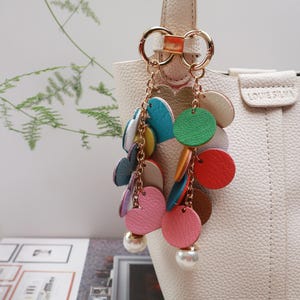 May include: A white handbag with a gold chain and colorful leather charms. The charms are in the shape of circles and hearts. The bag has a gold clasp and a gold handle.
