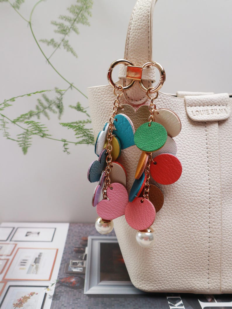 May include: A white handbag with a gold chain and colorful leather charms. The charms are in the shape of circles and hearts. The bag has a gold clasp and a gold handle.