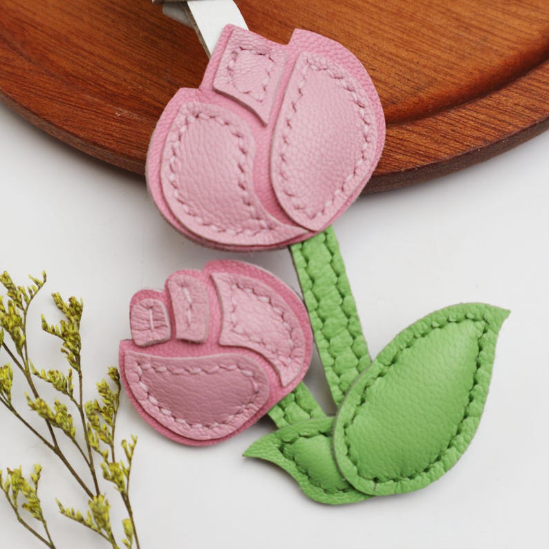May include: A leather keychain featuring a pink tulip design with green leaves and stems. The tulip petals are a soft pink, and the leaves are a vibrant green. The keychain is likely handmade and has a stitched design.