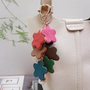 May include: A white handbag with a gold chain and a colorful star-shaped keychain. The keychain has five stars in different colors: red, pink, brown, green, and blue. The stars are made of leather and have a pearl-like bead at the bottom.