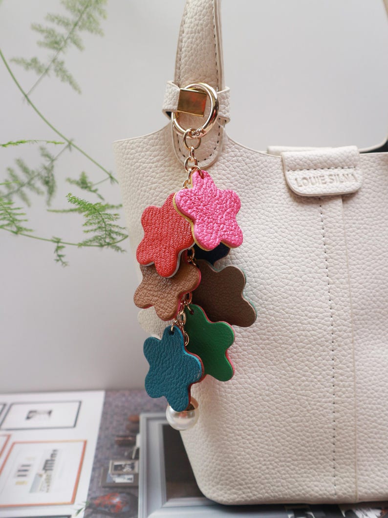 May include: A white handbag with a gold chain and a colorful star-shaped keychain. The keychain has five stars in different colors: red, pink, brown, green, and blue. The stars are made of leather and have a pearl-like bead at the bottom.