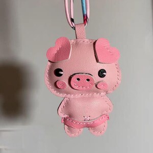 May include: A pink pig keychain made of leather with stitched details. The pig has heart-shaped ears, black eyes, and a pink snout.
