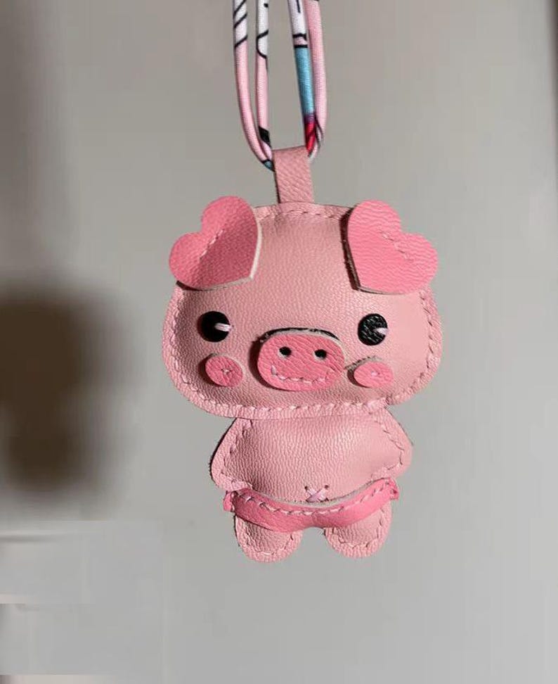 May include: A pink pig keychain made of leather with stitched details. The pig has heart-shaped ears, black eyes, and a pink snout.