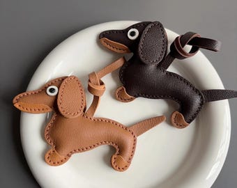 May include: Two leather dachshund keyrings. One is light brown, the other is dark brown. Both have white eyes and a loop for attaching to keys. The keyrings are on a white plate.