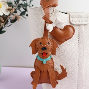 May include: A brown leather dog-shaped bag charm with a blue collar and red tongue, attached to a white handbag. A brown heart and white bow accent the bag. The handbag has the text "LOUIE SILNA".