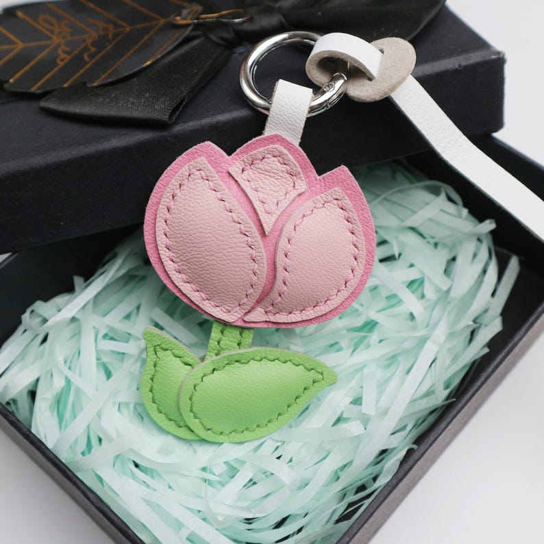 May include: A pink and light pink leather tulip keychain with green leaves, attached to a silver keyring and white leather strap. The keychain is presented in a black gift box with light blue shredded paper.