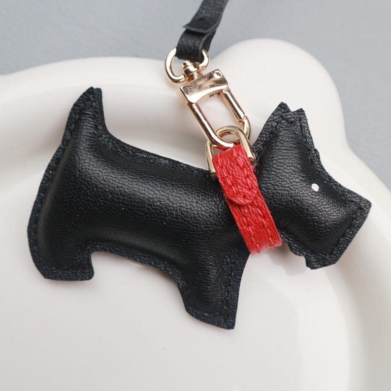 May include: A black leather Scottie dog keychain with a red collar and gold-tone clasp. The dog has a white dot for an eye and a black strap. This accessory is a charming addition to any set of keys.