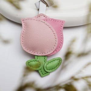 May include: A leather tulip-shaped keychain in shades of pink and green. The flower has two pink petals with stitched edges, green leaves, and a white leather strap for attachment. The design is detailed and handcrafted.