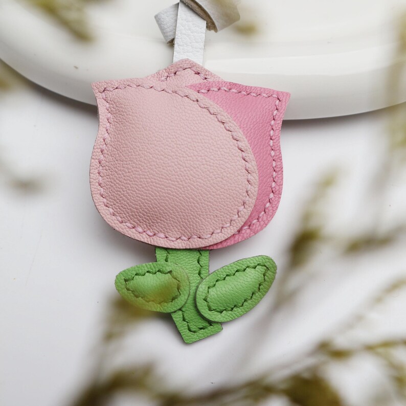 May include: A leather tulip-shaped keychain in shades of pink and green. The flower has two pink petals with stitched edges, green leaves, and a white leather strap for attachment. The design is detailed and handcrafted.