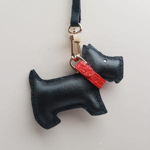 May include: A black leather dog-shaped keychain with a red collar. The keychain has a gold-colored clasp and a black strap. The dog's silhouette is detailed with stitched edges, creating a charming accessory.
