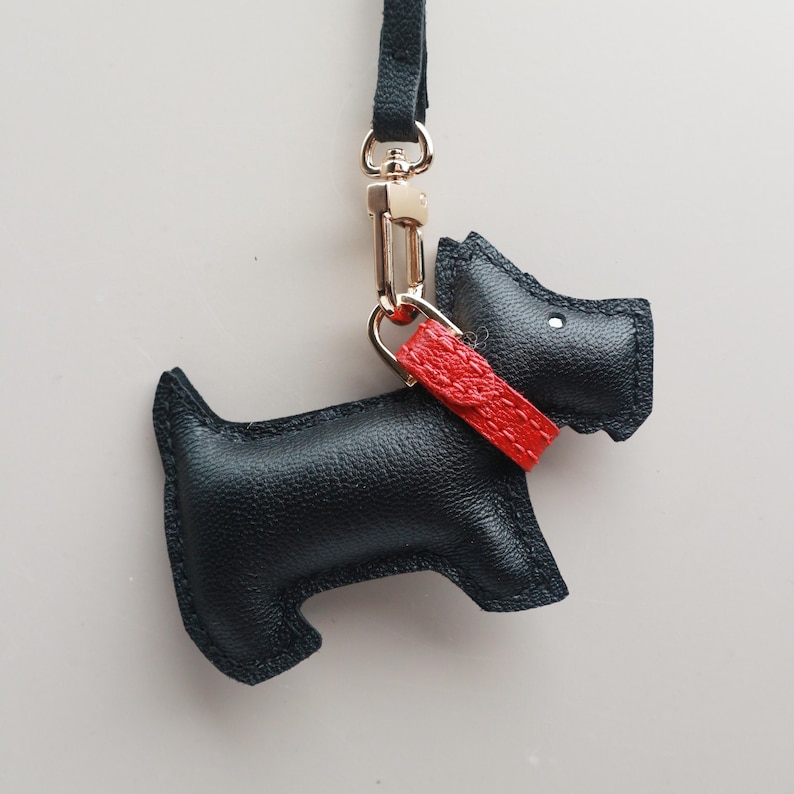 May include: A black leather dog-shaped keychain with a red collar. The keychain has a gold-colored clasp and a black strap. The dog's silhouette is detailed with stitched edges, creating a charming accessory.