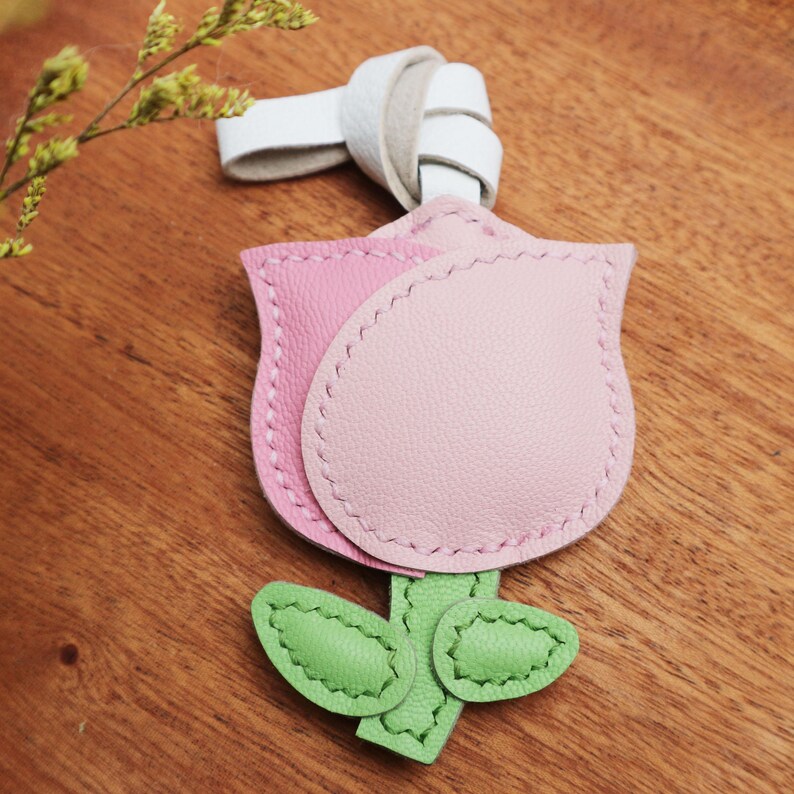 May include: A pink and light pink leather tulip-shaped keychain with green leaves and stem. The keychain has a white leather loop at the top. The tulip is stitched with white thread.