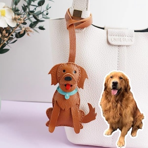 May include: A brown leather dog-shaped bag charm with a blue collar and red tongue, attached to a white handbag. A sticker of a golden retriever is next to the charm. The bag charm is a decorative accessory.
