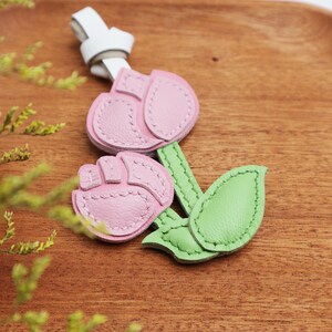 May include: A leather flower keychain with pink petals, a green stem and leaf, and a white strap. The flower is made of stitched leather and has a detailed design. The keychain is on a wooden surface.