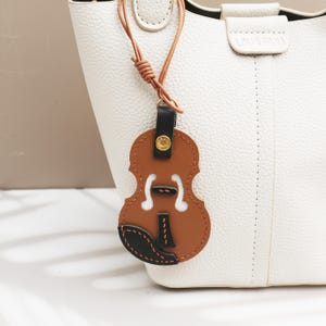 Puede incluir: Un charm de bolso de cuero marrón y negro con forma de violín y un botón dorado. El charm cuelga de una correa de cuero marrón claro. El fondo es un bolso de cuero blanco.