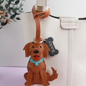 May include: A brown leather dog-shaped keychain with a blue collar and a red tongue. A black bone-shaped tag with the name "Rocky" is attached. The keychain hangs from a brown leather strap on a white leather bag.