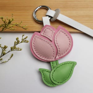 May include: A pink and light pink leather tulip keychain with green leaves. The flower is stitched with a darker pink thread. The keychain has a silver ring and a white leather strap.