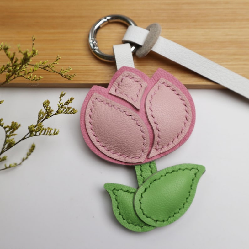 May include: A pink and light pink leather tulip keychain with green leaves. The flower is stitched with a darker pink thread. The keychain has a silver ring and a white leather strap.