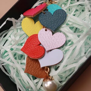 May include: A colorful leather keychain with a pearl charm. The keychain features seven heart-shaped charms in different colors, including red, pink, yellow, green, blue, brown, and white.