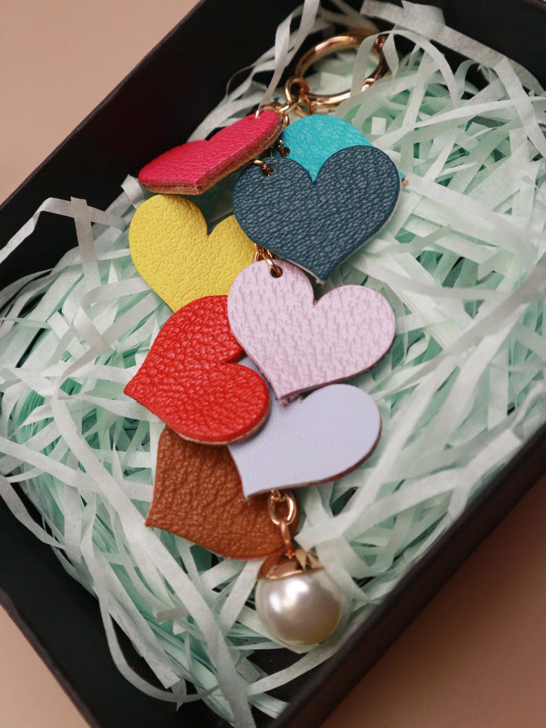 May include: A colorful leather keychain with a pearl charm. The keychain features seven heart-shaped charms in different colors, including red, pink, yellow, green, blue, brown, and white.