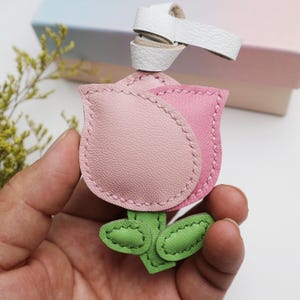 May include: A pink leather flower-shaped keychain with green leaves and a white leather loop. The flower has a stitched outline and a light pink hue. The leaves are a bright green color.
