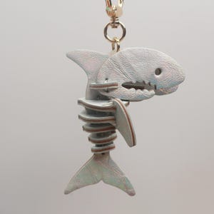 May include: A shark-shaped keychain with a skeleton design. The keychain is made of iridescent, textured material with a gold-colored clasp. The shark's body is light gray with brown accents, and the design includes a tail, fins, and a jaw.