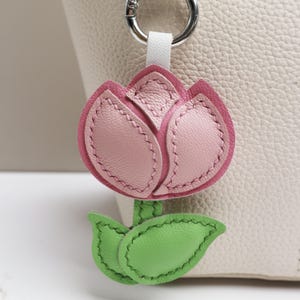 May include: A pink and green leather tulip keychain with a silver-tone clasp. The tulip has pink petals with darker pink edging and green leaves. The keychain is attached to a white leather strap.