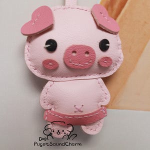 May include: A pink leather pig keychain with heart-shaped ears, black eyes, and a pink snout. The keychain is stitched with pink thread and has a pink leather strap.