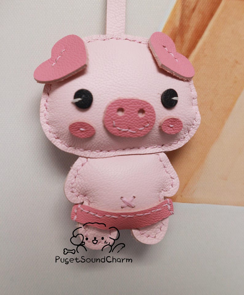 May include: A pink leather pig keychain with heart-shaped ears, black eyes, and a pink snout. The keychain is stitched with pink thread and has a pink leather strap.