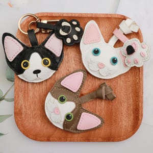 Può includere: Portachiavi a forma di gatto fatti a mano in pelle nera, bianca e marrone. Ogni portachiavi presenta un design a forma di muso di gatto con dettagli rosa e hardware color oro. I portachiavi sono esposti su un vassoio di legno.