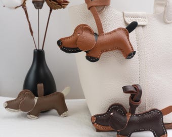 May include: Three brown leather dachshund dog keychains. The keychains are hanging from a white leather bag. The keychains are stitched with brown thread.