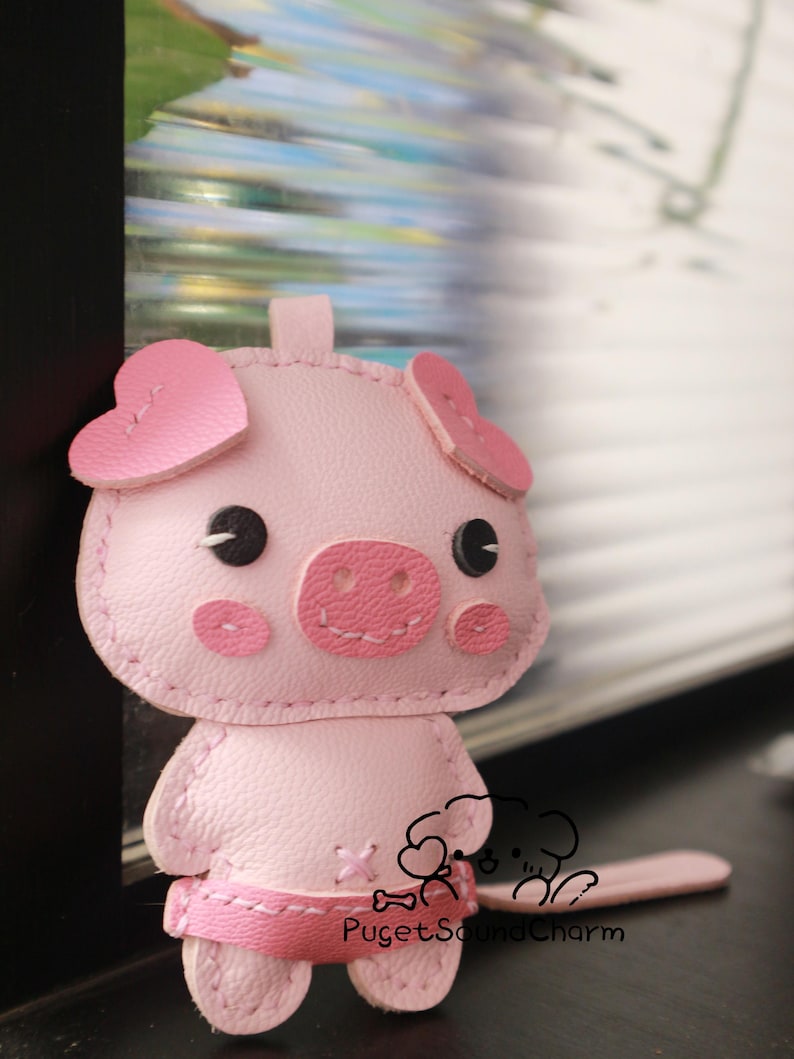 May include: A pink leather pig keychain with heart-shaped ears and a stitched-on smile. The keychain has a small, pink leather tail and the text "Puget Sound Charm" is stitched on the bottom.