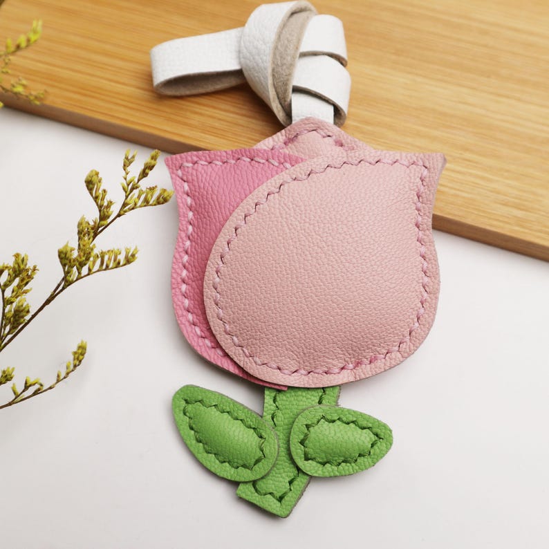 May include: A pink and light pink leather tulip-shaped keychain with green leaves and a white leather loop. The keychain is stitched with a contrasting thread and is set against a light wooden surface.