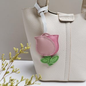 May include: A cream-colored leather handbag with a pink and light pink leather tulip-shaped charm attached. The charm has green leaves and a white leather strap. The handbag has the text "LOUIE SILMA" on the front.